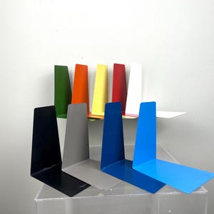 May include: A selection of brightly coloured metal bookends in shades of green, orange, yellow, red, white, black, grey, and blue. The L-shaped bookends are designed to support books. The bookends are displayed on a clear surface.
