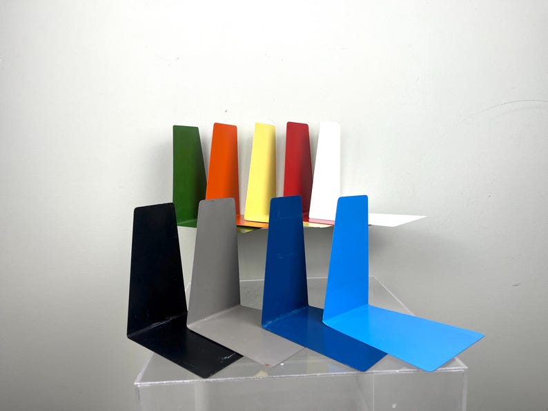 May include: A selection of brightly coloured metal bookends in shades of green, orange, yellow, red, white, black, grey, and blue. The L-shaped bookends are designed to support books. The bookends are displayed on a clear surface.