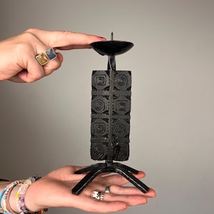 May include: A black metal candle holder featuring a central rectangular column decorated with carved spiral patterns on all sides. The piece includes a circular top plate with a central spike for a candle and a tripod base with three legs.