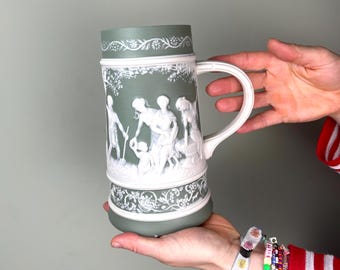 May include: A green and white ceramic mug with a handle. The mug features a relief design of figures in a pastoral scene. The handle and decorative trim are white, while the body of the mug is green. The mug is held in a person's hands.