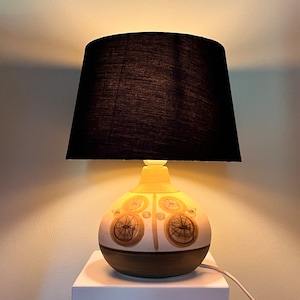 Könnte beinhalten: Eine Keramik-Tischlampe mit schwarzem Lampenschirm. Der Lampenfuß ist cremefarben mit braunen und beigen kreisförmigen Designs. Die Lampe steht auf einem weißen Sockel, und das Kabel ist sichtbar.