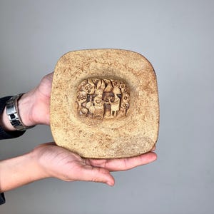 May include: A square, tan-coloured ceramic dish with rounded corners and a textured, speckled surface. The centre of the dish features a recessed, circular relief carving depicting a group of stylised figures and circular patterns in a monochromatic tone.