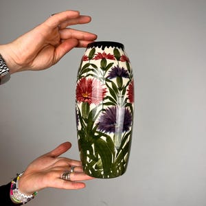 Könnte beinhalten: Eine zylindrische Keramikvase mit cremefarbener Basis, verziert mit einem botanischen Muster aus roten und violetten Blumen, grünen Blättern und Stängeln. Die Vase hat eine glatte, glänzende Oberfläche und einen schwarzen Rand.