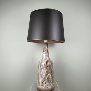 Könnte beinhalten: Eine Tischlampe aus Keramik mit einem dunkelgrauen Lampenschirm. Der Lampenfuß hat eine strukturierte, getropfte Glasur in Braun-, Beige- und Weißtönen. Die Lampe ist an eine Steckdose angeschlossen. Die Lampe steht auf einem transparenten Acrylständer.