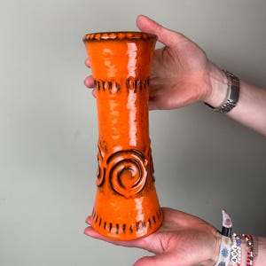Op de afbeelding: Een hoge, oranje keramische vaas met een glanzende afwerking. De vaas heeft een uitlopende basis, een cilindrisch lichaam met spiraal- en verticale lijnen en een getextureerde rand. De vaas is ongeveer 25 cm hoog.