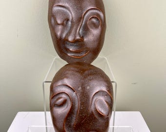 Könnte beinhalten: Zwei braune Keramikmasken mit ausdrucksstarken Gesichtern, eine lächelnd und eine mit einem ernsteren Ausdruck. Die Masken werden auf einem klaren Acrylständer präsentiert.