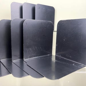 May include: A set of black metal bookends. The bookends are L-shaped with a rounded top and a flat base. The bookends are arranged in a row, with some standing upright and others lying flat. The bookends are made of a dark, matte metal.