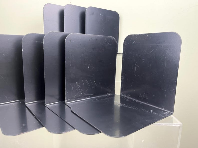 May include: A set of black metal bookends. The bookends are L-shaped with a rounded top and a flat base. The bookends are arranged in a row, with some standing upright and others lying flat. The bookends are made of a dark, matte metal.