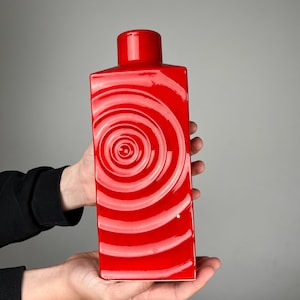 Peut inclure: Un vase rectangulaire en céramique rouge avec un dessus cylindrique. Le vase présente un motif en spirale blanc sur le devant. Le vase est tenu par deux mains sur un fond gris. Le vase mesure environ 25 cm de haut.