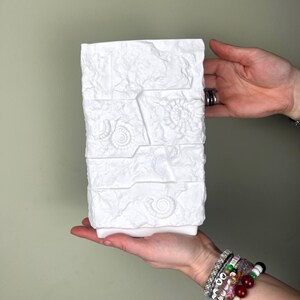 May include: A white rectangular vase with a textured, stone-like appearance. The vase features embossed seashell designs and a rectangular base. The vase is held in a person's hands against a neutral background.
