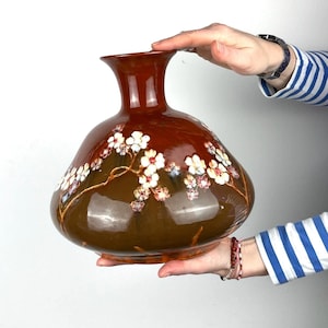 Könnte beinhalten: Eine große, braune Keramikvase mit einem breiten, abgerundeten Boden und einem schmalen Hals. Die Vase ist mit einem floralen Muster aus weißen Blüten mit rosa und gelben Akzenten verziert.