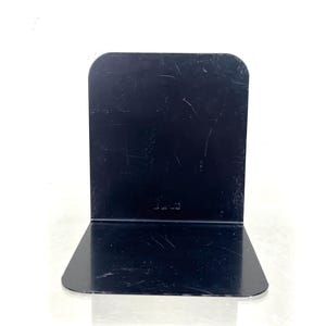 May include: A black metal bookend with a rectangular back and a right-angled base. The bookend has a glossy finish and a few scratches. The word "Luce" is etched on the back. The bookend is designed to hold books upright.