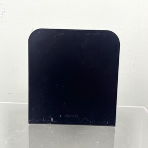 May include: A black, rectangular object with rounded top corners. It stands upright on a reflective surface. The background is a plain white wall.