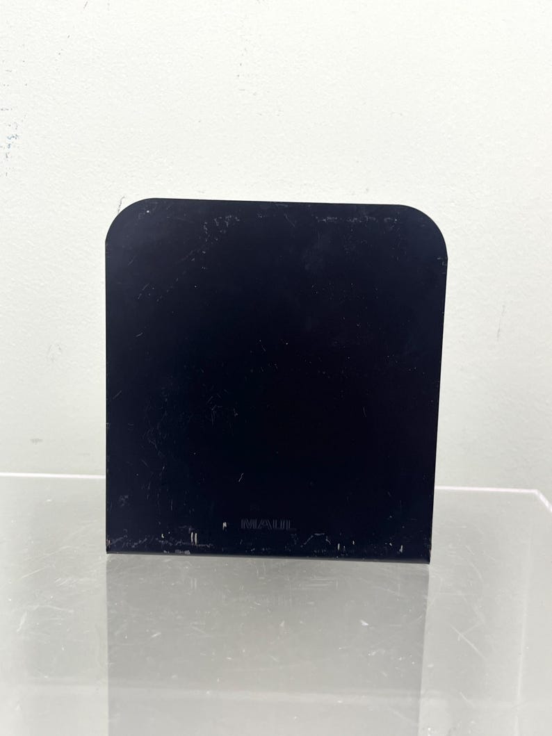May include: A black, rectangular object with rounded top corners. It stands upright on a reflective surface. The background is a plain white wall.