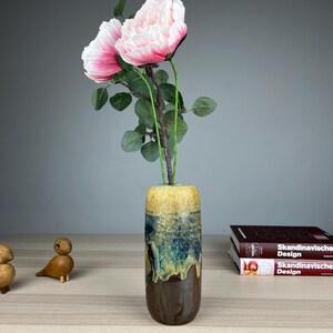 Könnte beinhalten: Eine Keramikvase mit Farbverlauf, die rosa und weiße Kunstblumen enthält. Die Vase geht von Braun zu Blau und Gelb über. Zwei kleine Holzvogelfiguren stehen auf dem Tisch.