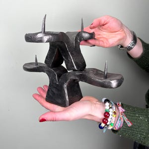 Könnte beinhalten: Eine dunkelgraue, abstrakte Metallskulptur mit drei nach oben gerichteten Spitzen. Die Skulptur hat ein geschichtetes Design mit zwei Plattformen, die durch einen zentralen Träger verbunden sind. Das Stück wird von zwei Händen gehalten.