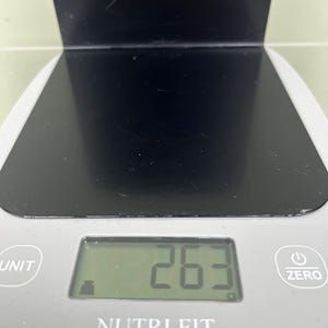 May include: A digital kitchen scale with a black surface and a grey base. The display shows the number 263. The scale has buttons labelled "UNIT" and "ZERO", and the brand name "NUTRI FIT" is visible.