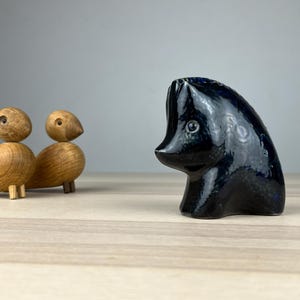 Könnte beinhalten: Eine dunkelblaue Keramikskulptur eines stilisierten Tierkopfes mit glänzender Oberfläche. Zwei Holzfiguren von Vögeln mit runden Körpern und kleinen Beinen befinden sich links von der Skulptur. Der Hintergrund ist hellgrau.