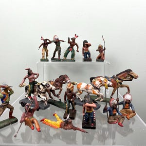 May include: A collection of vintage toy figurines depicting Native American figures and horses. The figurines are painted in various colours and poses, including warriors, riders, and figures with bows and arrows. The collection is displayed on a clear, multi-tiered shelf.