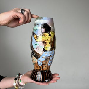 Pode incluir: Um vaso decorativo com uma cena pintada de uma pessoa com um casaco amarelo e calças azuis. O vaso tem uma base preta com listras verticais e um topo alargado. A obra de arte apresenta uma variedade de cores, incluindo azul, amarelo e castanho.