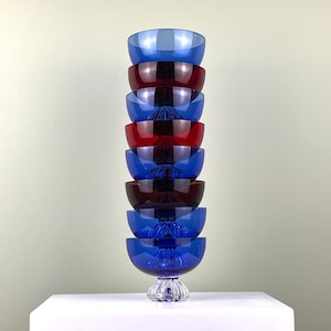 May include: A stack of eight glass bowls, alternating between blue and red, with a clear glass base. The bowls are stacked on a white surface.