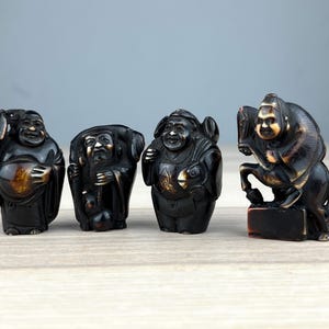 May include: Four small, dark-coloured figurines of smiling figures. The figures are carved with intricate details, and each has a unique pose and expression. The figurines are displayed on a light-coloured wooden surface.