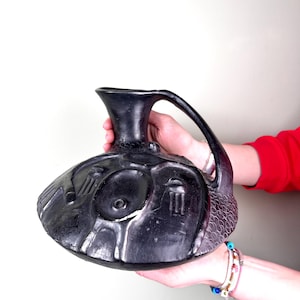 Puede incluir: Una jarra de cerámica negra con una base ancha y redondeada y un asa. La jarra tiene intrincados tallados y una superficie texturizada.