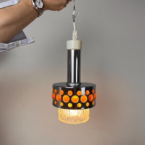 May include: A pendant light fixture featuring a cylindrical chrome body with a band of circular orange glass inserts. Below the chrome section is a clear, textured glass diffuser. The fixture includes a white plastic top cap and cord.