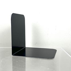 May include: A single, black metal bookend with a right-angle design. The bookend has a rectangular base and a tall, vertical support. The surface is smooth and reflective, and the bookend is positioned on a clear surface against a white background.