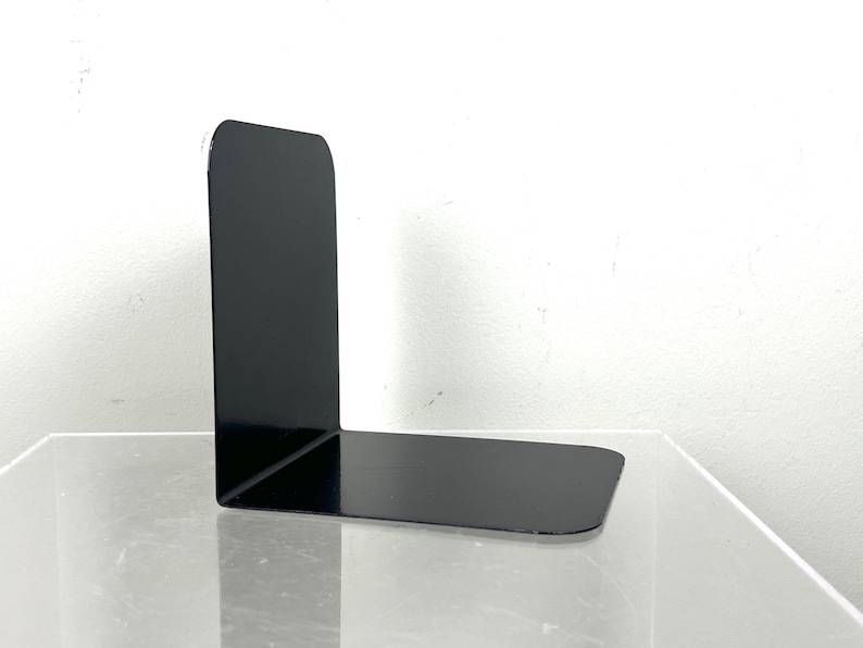 May include: A single, black metal bookend with a right-angle design. The bookend has a rectangular base and a tall, vertical support. The surface is smooth and reflective, and the bookend is positioned on a clear surface against a white background.