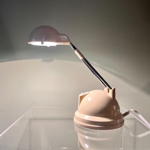 Könnte beinhalten: Eine weiße Schreibtischlampe mit einem runden, kuppelförmigen Schirm und einem flexiblen Arm. Die Lampe steht auf einem weißen Sockel mit einer runden, kuppelförmigen Oberseite.