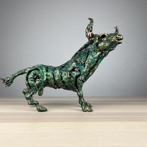May include: A bronze sculpture of a bull in a dynamic pose, with a green and gold patina. The bull's body has an open, cage-like design, and its horns are curved upwards. The sculpture is on a light-colored surface.