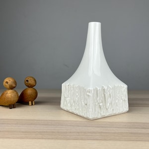 May include: A white ceramic vase with a textured, bark-like design on the base and a smooth, flared top. Two small, wooden bird figurines are to the left of the vase, adding a touch of natural decor.