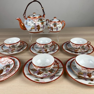 Pode incluir: Um conjunto de chá de estilo japonês com bule, açucareiro, xícaras e pires. O conjunto de porcelana branca é decorado com uma cena de paisagem e figuras, com detalhes em vermelho e dourado. O conjunto inclui seis xícaras e pires e duas travessas.