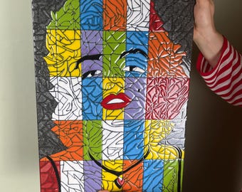 May include: A colorful mosaic art piece depicting a portrait of Marilyn Monroe. The artwork is divided into square sections, each with a different color, including red, yellow, green, and purple. The background is a textured gray.