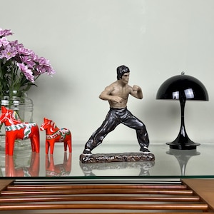 May include: A figurine of a martial artist in a fighting stance, wearing dark trousers, positioned next to two red painted wooden horse figurines and a black dome-shaped lamp. A glass vase containing pink flowers is also present on the surface.