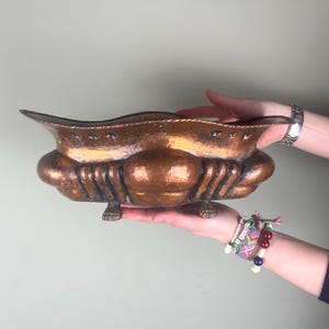 May include: A hammered copper decorative bowl with a scalloped rim and ornate, rounded sides. The bowl is supported by small feet. The bowl is held by two hands.