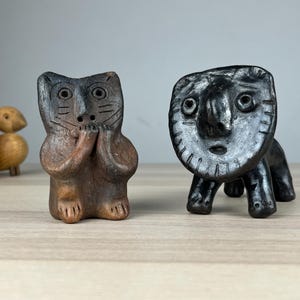 Puede incluir: Dos figuras de cerámica hechas a mano. Una es una figura oscura con forma de gato con las patas cerca de su cara. La otra es una criatura negra estilizada con una cara grande y cuatro patas. Figuras de patos de madera en el fondo.