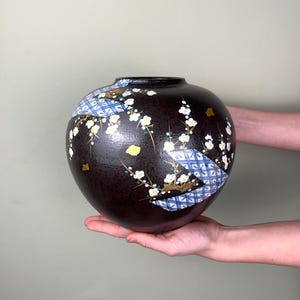 Può includere: Un vaso rotondo in ceramica marrone scuro con un collo stretto. Il vaso è decorato con rami floreali bianchi, accenti in foglia d'oro e un motivo geometrico blu e bianco. Il vaso è tenuto tra le mani di una persona.