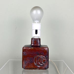 Könnte beinhalten: Eine Vintage-Tischlampe mit quadratischem, rotbraunem Keramiksockel. Der Sockel hat ein kreisförmiges Design und trägt eine weiße Lampenfassung und eine mattierte Glühbirne. Die Lampe steht auf einer transparenten Oberfläche.