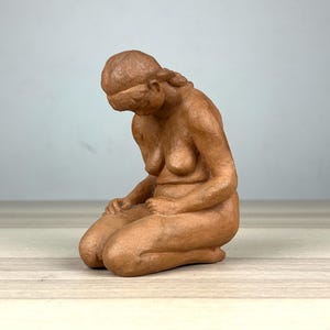 Könnte beinhalten: Braune Keramikskulptur einer knienden Figur mit gesenktem Kopf. Die Figur sitzt mit angewinkelten Knien und Händen auf den Oberschenkeln. Die Skulptur hat eine glatte, matte Oberfläche und steht vor einem neutralen Hintergrund.