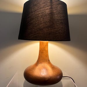 Può includere: Una lampada da tavolo in legno con un paralume marrone scuro. La base della lampada è di colore marrone caldo con un fondo arrotondato e un collo sottile. Il paralume è in tessuto marrone scuro e la lampada è collegata.