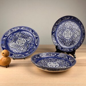 May include: Three ceramic plates featuring a deep blue and white intricate floral and geometric pattern. Two plates are displayed upright on black stands, while one plate rests flat. A small wooden bird figurine is positioned to the left.