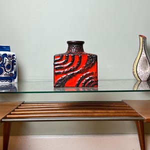 Puede incluir: Tres jarrones decorativos de cerámica sobre una estantería de cristal. Los jarrones incluyen un jarrón cilíndrico con estampado blanco y azul, un jarrón cuadrado con textura roja y negra y un jarrón alto y delgado con diseño amarillo y negro.