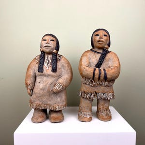 Könnte beinhalten: Zwei Keramikfiguren von amerikanischen Ureinwohnern, beide mit langen schwarzen Zöpfen, in traditioneller Kleidung. Die Figur links ist eine Frau mit einem langen Kleid und die Figur rechts ist ein Mann mit einer kürzeren Tunika. Beide Figuren stehen auf einem weißen Sockel.