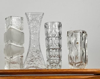 May include: Four clear glass vases with different textured designs. The vases are arranged in a row on a wooden surface.