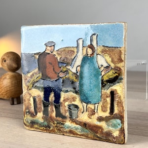 May include: A square ceramic tile featuring a hand-painted scene of two figures in a field. One figure wears a brown shirt and blue pants, while the other wears a blue apron. The background includes a light blue sky and earthy tones.