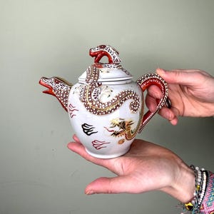 Könnte beinhalten: Weiße Keramik-Teekanne mit Drachendesign. Die Teekanne hat rote und goldene Akzente, mit einem drachenförmigen Ausguss und Griff. Der Deckel ist mit einer Drachenfigur gekrönt. Die Teekanne ist mit goldenen und schwarzen Illustrationen verziert.