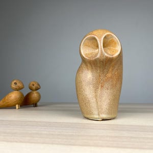 May include: A ceramic owl sculpture with a speckled tan and brown finish. Two small wooden bird figurines are to the left of the owl. The owl has large, open eye sockets and a rounded body. The background is a light gray.