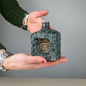 Könnte beinhalten: Kleine, quadratische Keramikvase mit strukturierter, teal-farbener Außenseite. Die Vase hat ein zentrales, vertieftes Design mit einer kupferfarbenen Spirale. Die Vase wird in den Händen einer Person gehalten, was ihre kompakte Größe und ihr einzigartiges Design hervorhebt.